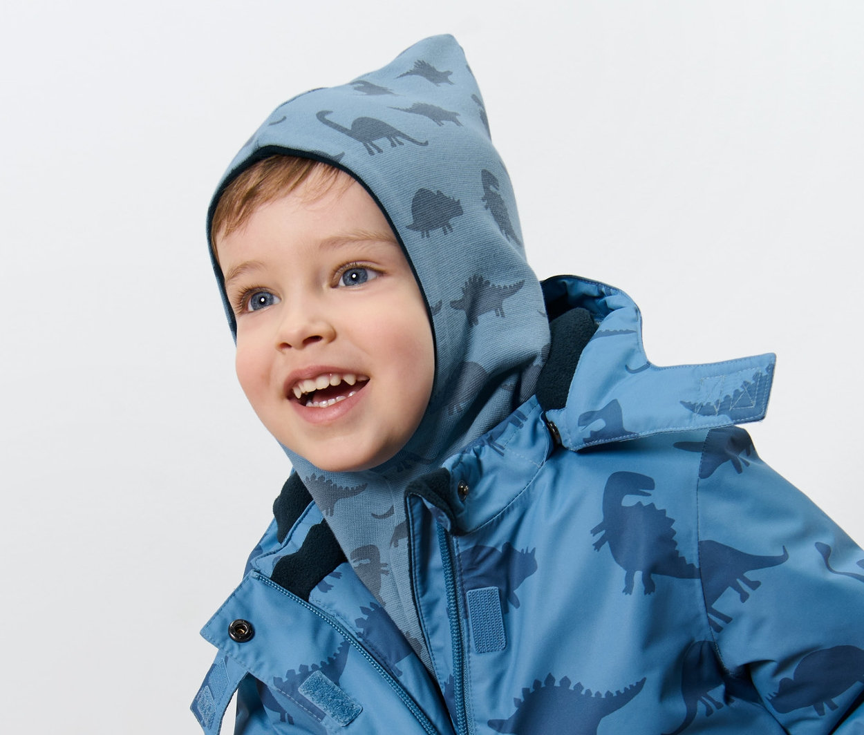 Egy fiú dinoszauruszos gyerek kifordítható belebújós sapkát és dinoszaurusz mintás kabátot visel.