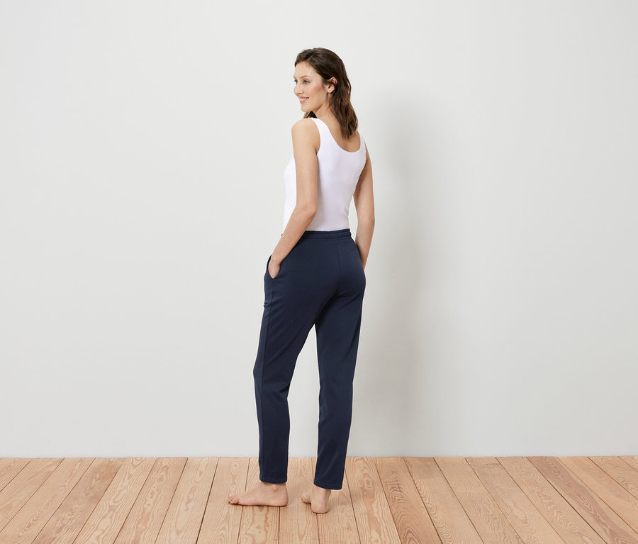 Nő fehér trikóban és sötétkék nadrágban áll háttal a kamerának, és a válla fölött néz hátra.