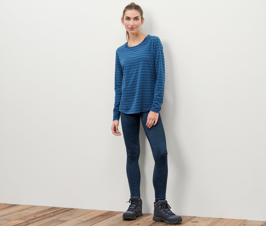 Nő áll, sportleggingset, kék csíkos hosszú ujjú inget és csizmát visel.