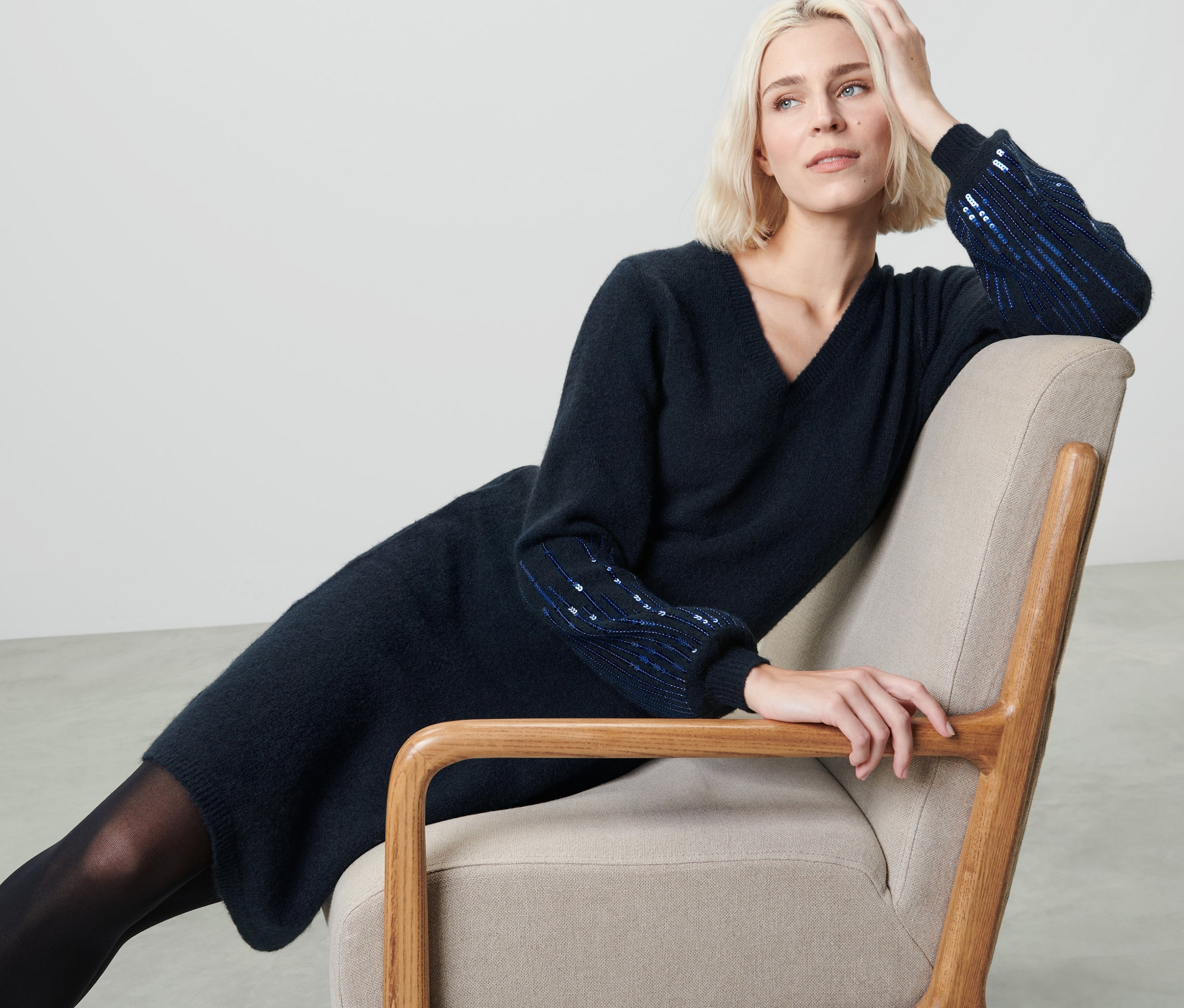 Szőke nő ül egy fotelben és sötétkék, flitteres kötött ruhát visel.