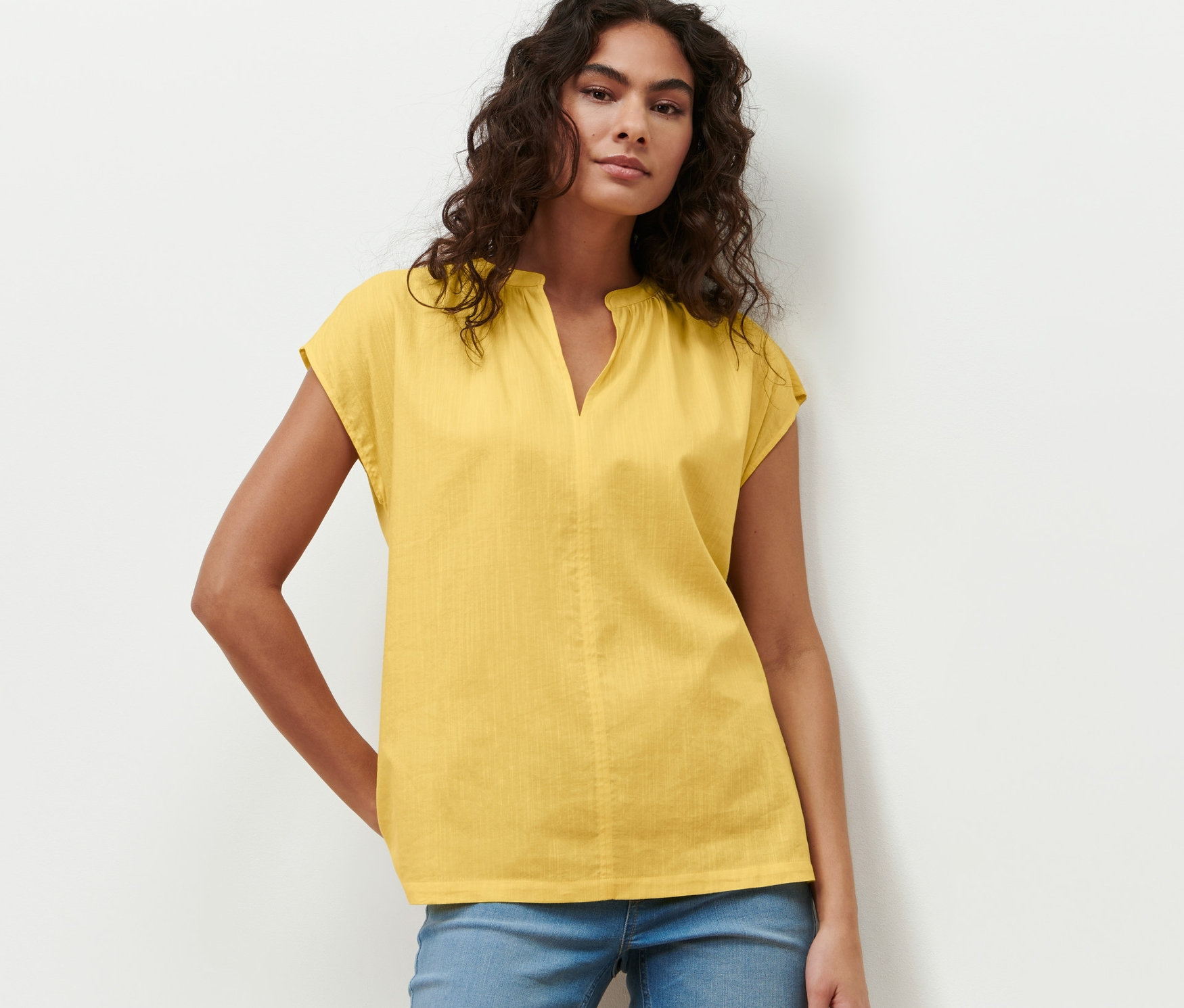 Nő pózol sárga rövid ujjú blúzban és kék farmerben.