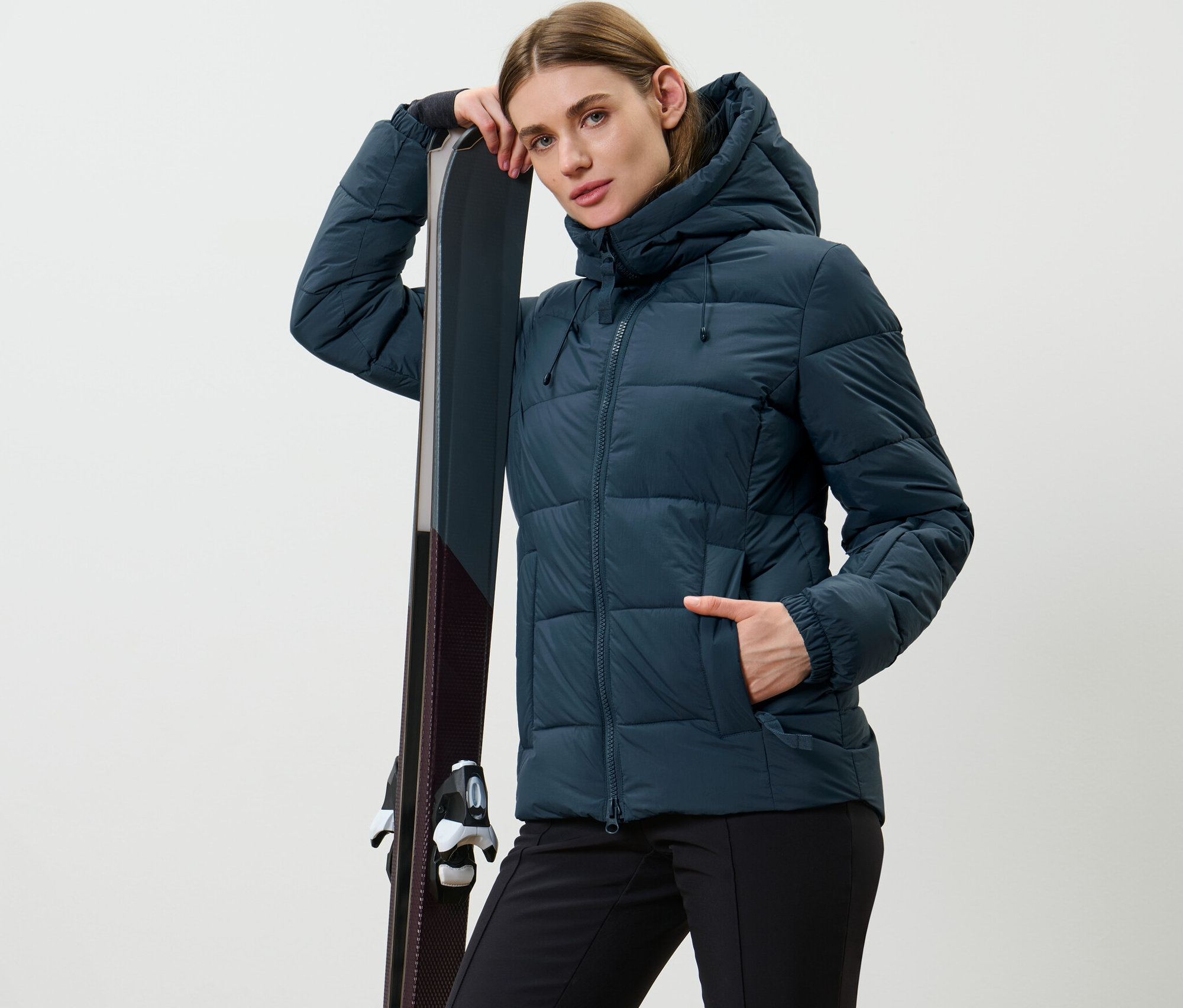 Nő pózol sötétkék steppelt síkabátban, és a sílécekre támaszkodik.