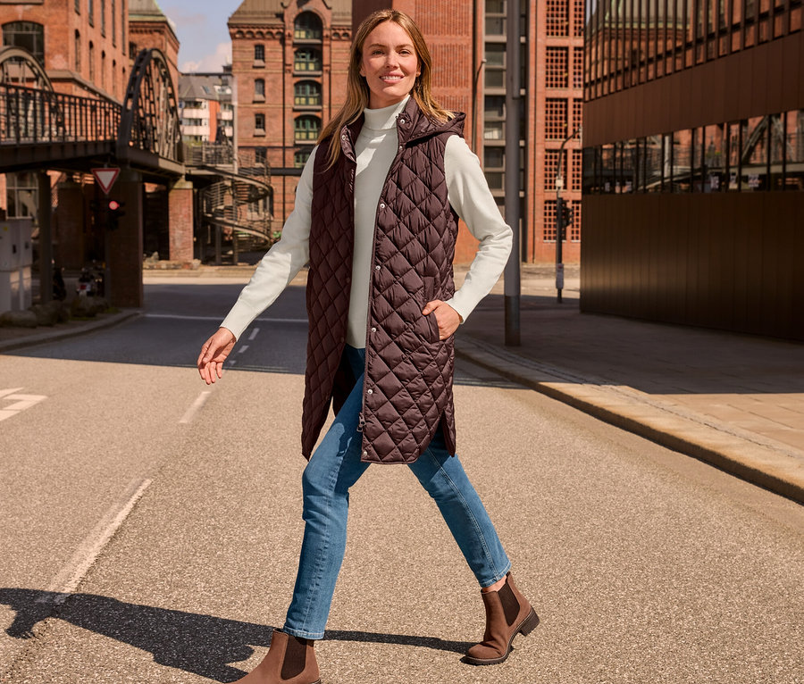 Nő sétál az utcán, hosszú steppelt mellény, krémfehér garbó, középkék farmernadrág és barna velúrbőr csizma van rajta.