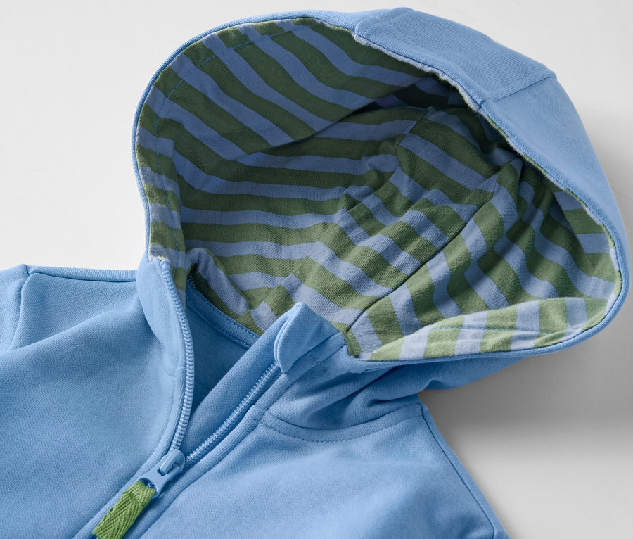 Egy világoskék, cipzáras kisfiú kapucnis szabadidőfelső részlete, kék-zöld csíkos belsővel.