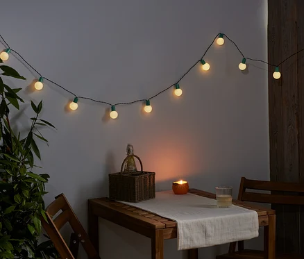 Egy LED fényfüzér lóg a falon. Az asztalon egy kosár, egy gyertya és egy pohár áll.