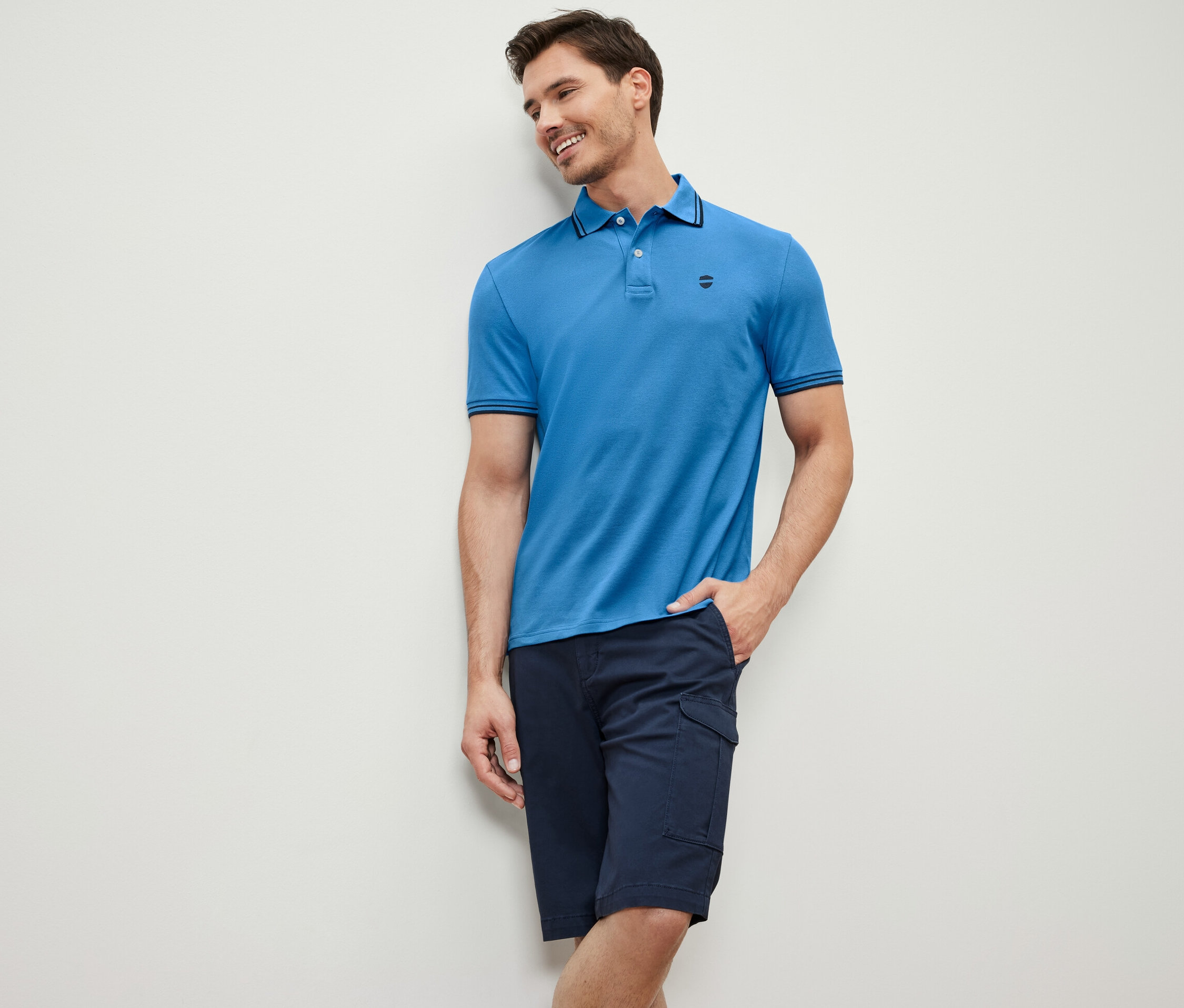 Egy falnak támaszkodó férfi kék piké pólót és sötétkék rövidnadrágot visel.