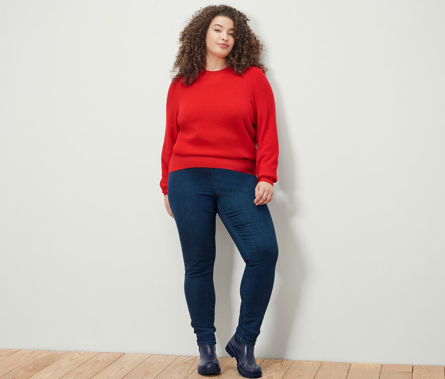 Nő pózol piros finomkötésű pulóverben és magasderekú farmer hatású leggingsben.