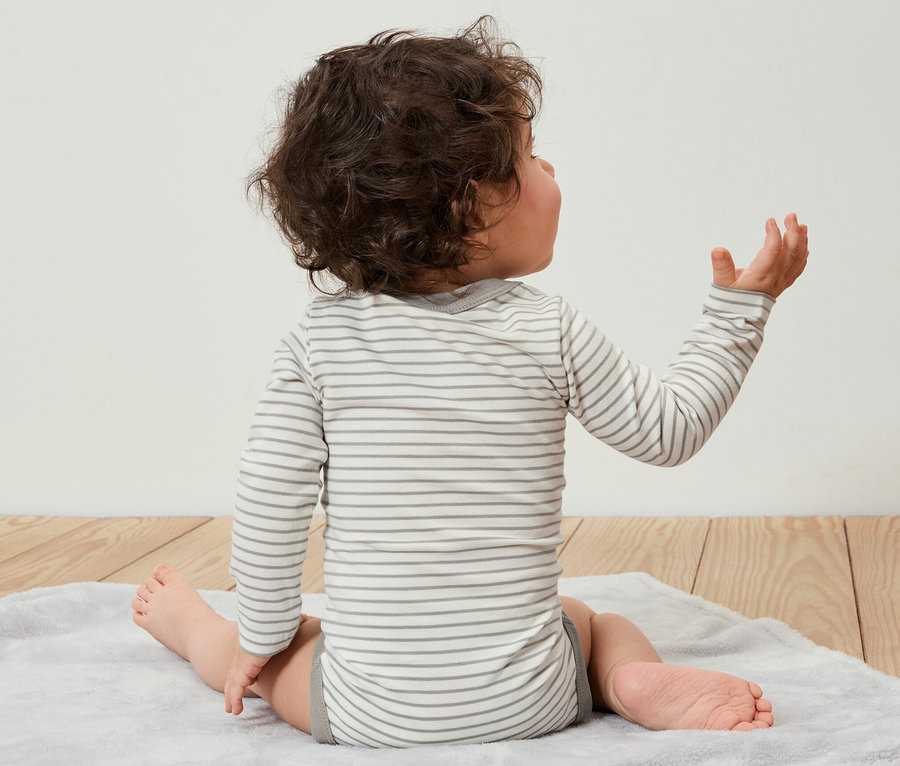 Egy hátulról látható kisgyerek ül egy szőnyegen, és 5 darabos, állítható hosszúságú bodyt visel.
