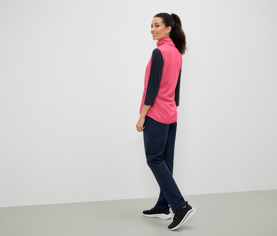 Egy nő a válla fölött mosolyog, rózsaszín mellényt, sötétkék nadrágot és fekete tornacipőt visel.