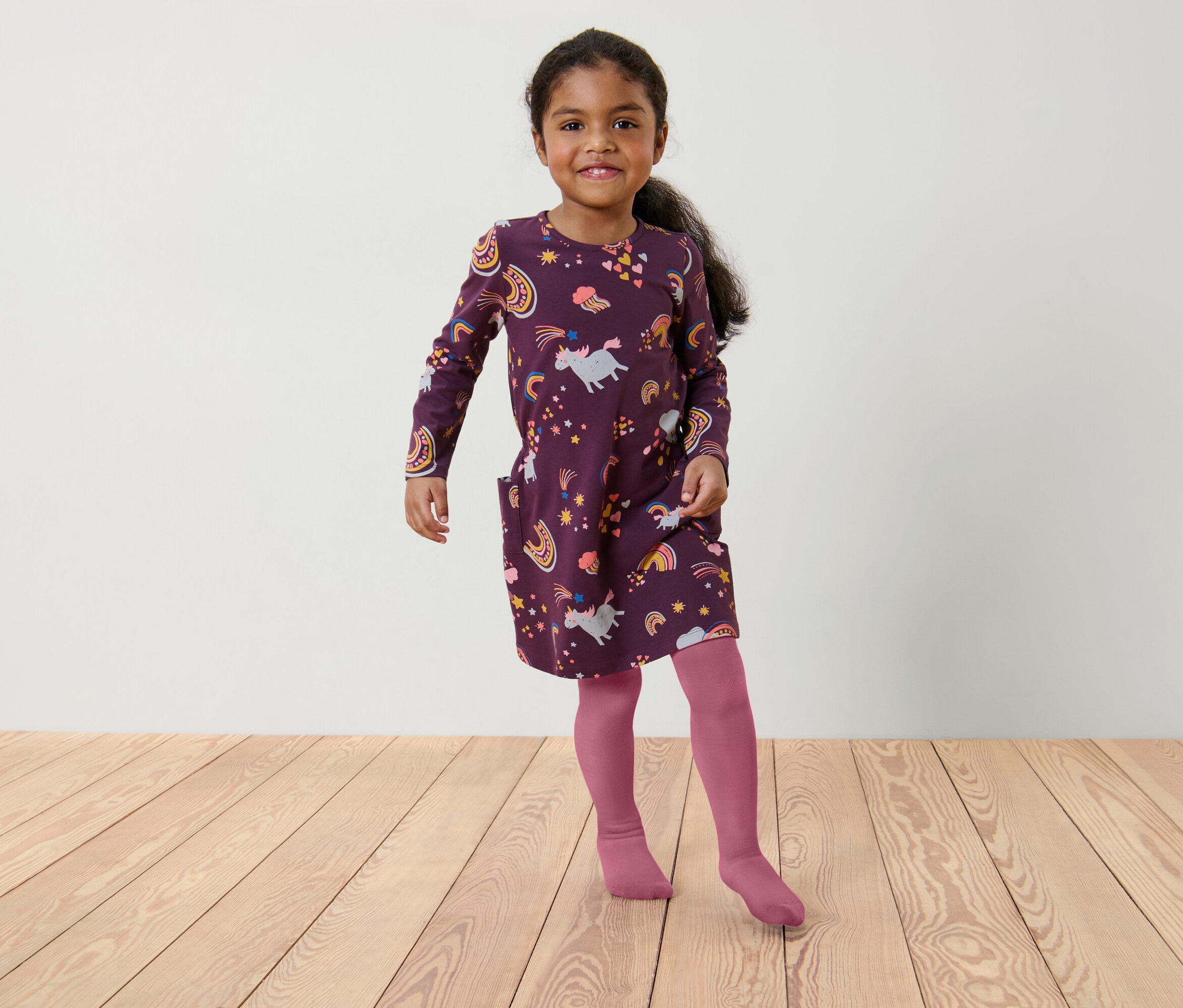 Egy kislány bordó, unikornis és szivárvány mintás ruhában és rózsaszín harisnyában áll egy fapadlón.