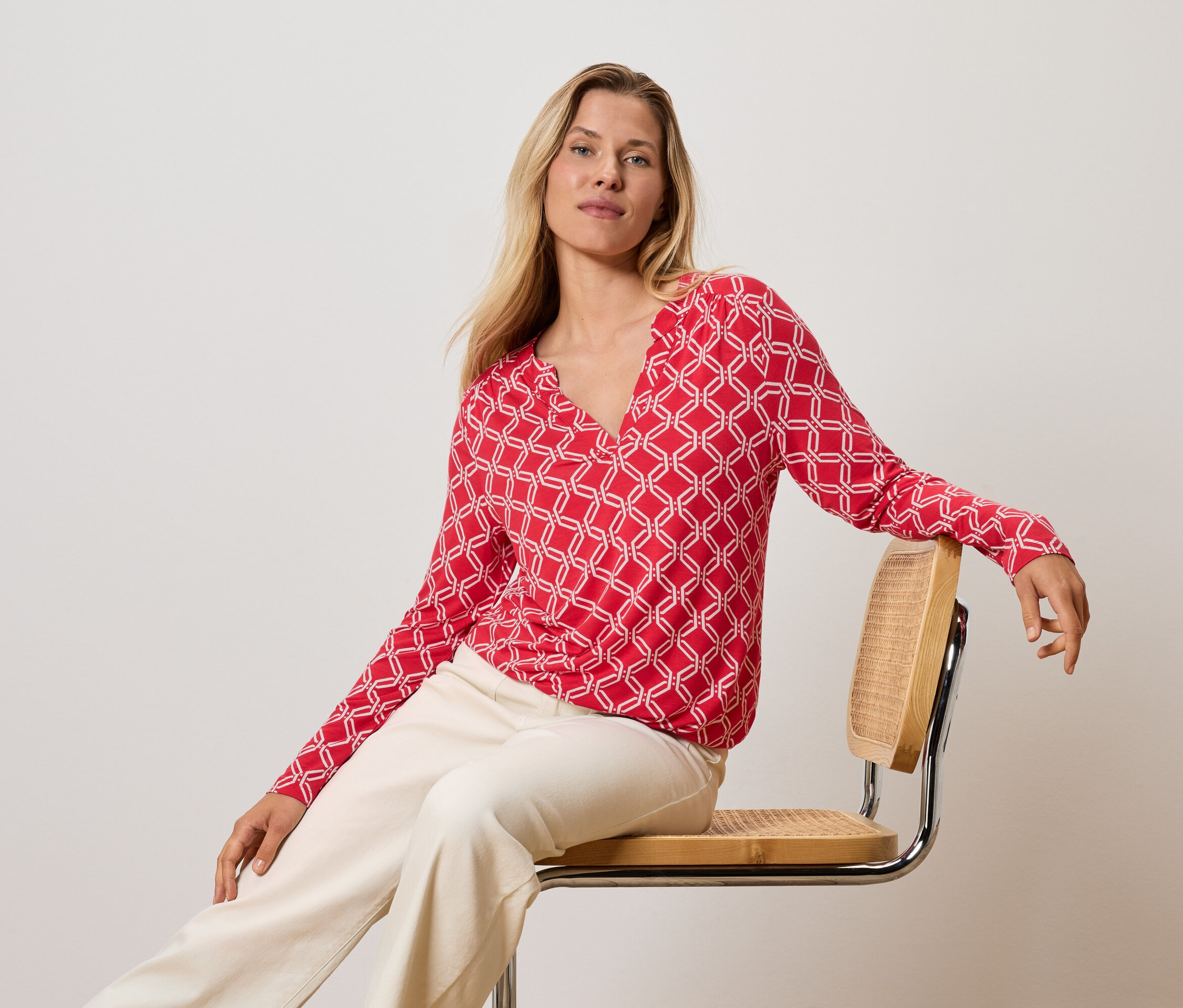 Szöszi nő ül egy széken, piros-fehér felsőben és krémszínű nadrágban.