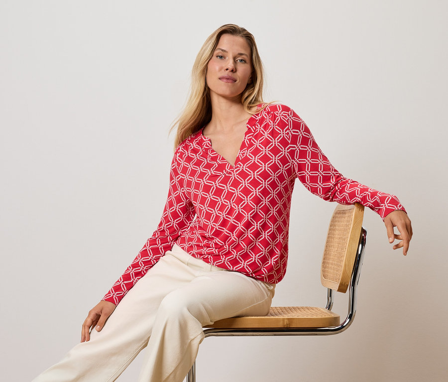 Szöszi nő ül egy széken, piros-fehér felsőben és krémszínű nadrágban.