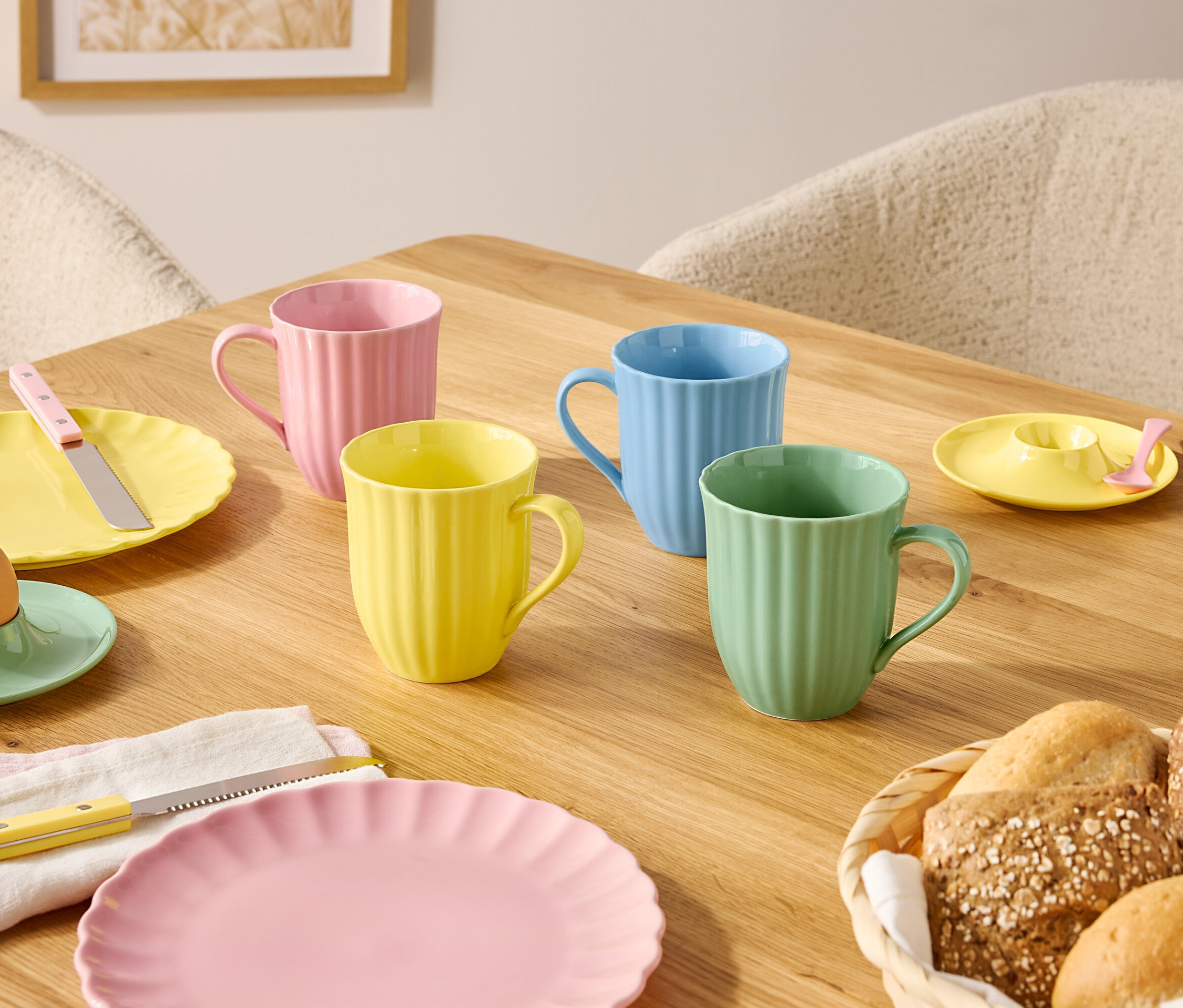 Egy fa asztalon pasztell színű tányérok és bögrék, valamint egy kenyérkosár található.