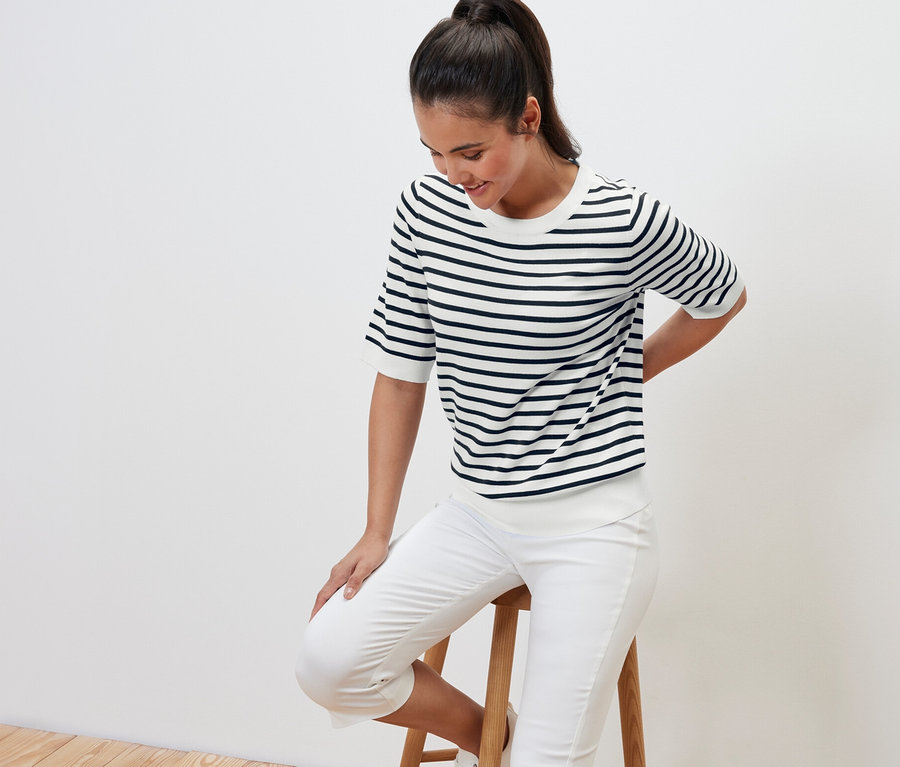 Nő ül egy sámlimon, tengerészkék, finom kötésű csíkos pulóvert visel.