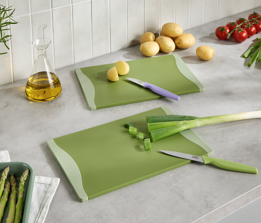 Két zöld vágódeszka vágott zöldségekkel, késekkel, burgonyával, paradicsommal, póréhagymával, spárgával és olajjal egy konyhapulton.