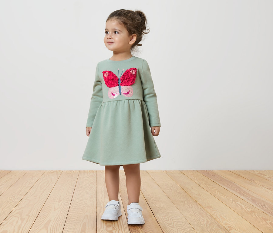 Kislány zöld pillangós ruhában áll a fapadlón.