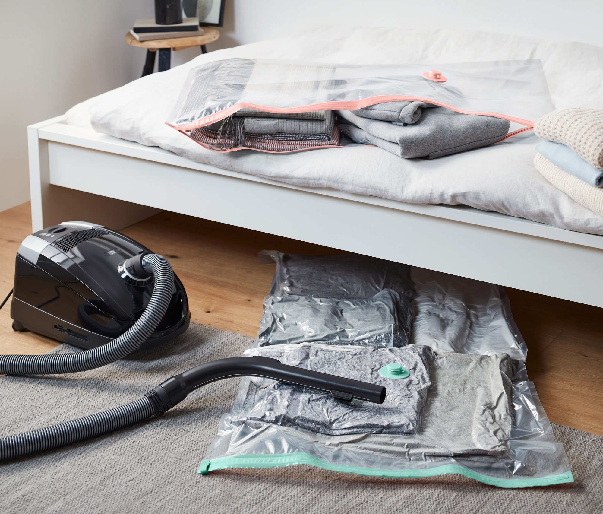 Porszívó fekszik két vákuumos tárolótasak mellett az ágy alatt. Egy vákuumos tárolótasak az ágyon fekszik.