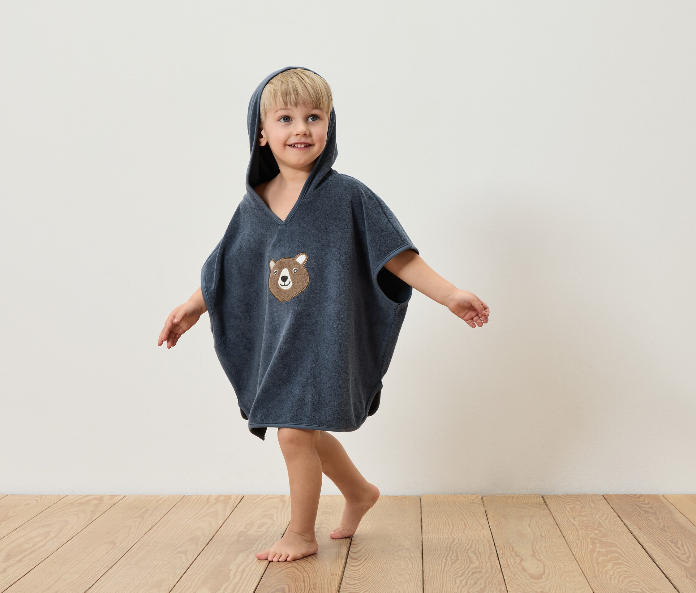 Egy kisfiú kék, kapucnis, medvemintás poncsót visel, és egy fa padlón áll egy fehér fal előtt.