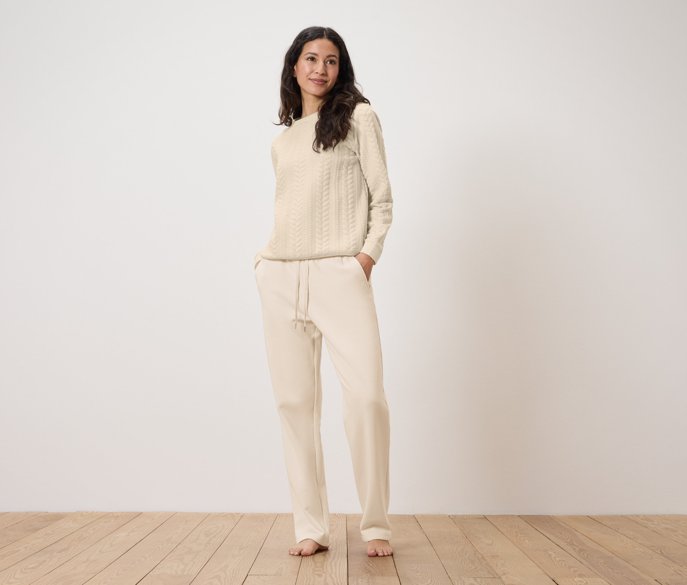 Egy nő áll, kezét a krémszínű, húzózsinóros joggingalsója zsebében tartja, hozzá krémszínű, kábelmintás kötött pulóvert visel.