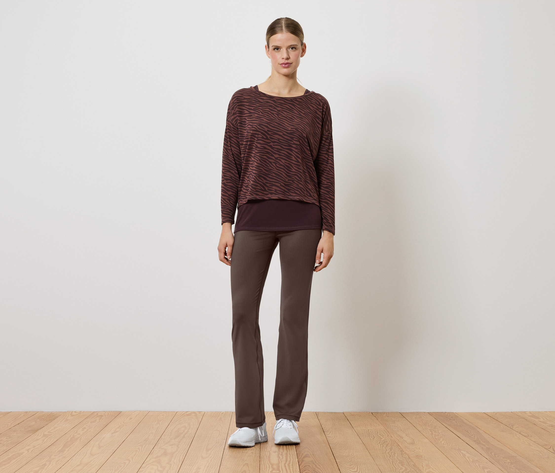 Egy nő barna pulóverben és nadrágban áll egy fapadlón.