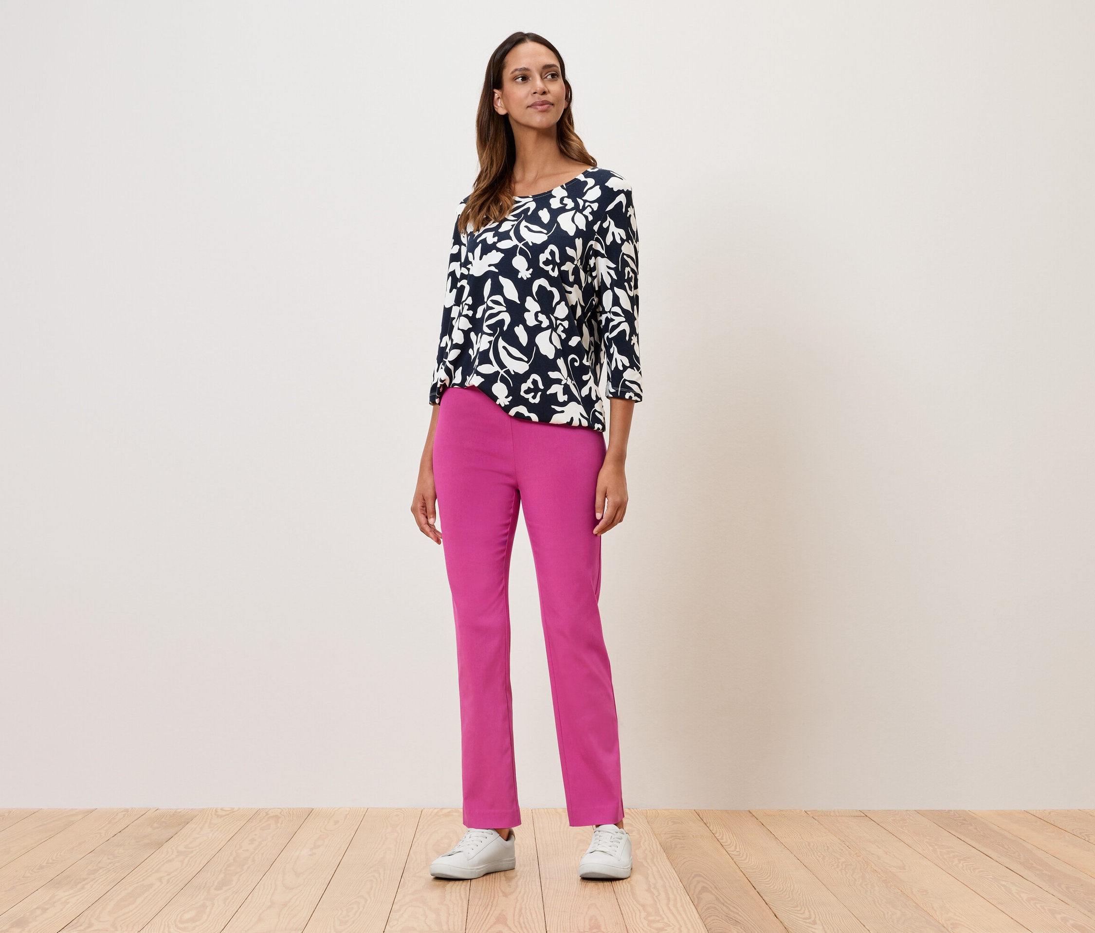 Egy nő áll virágos, háromnegyedes ujjú felsőben, rózsaszín nadrágban és fehér tornacipőben.