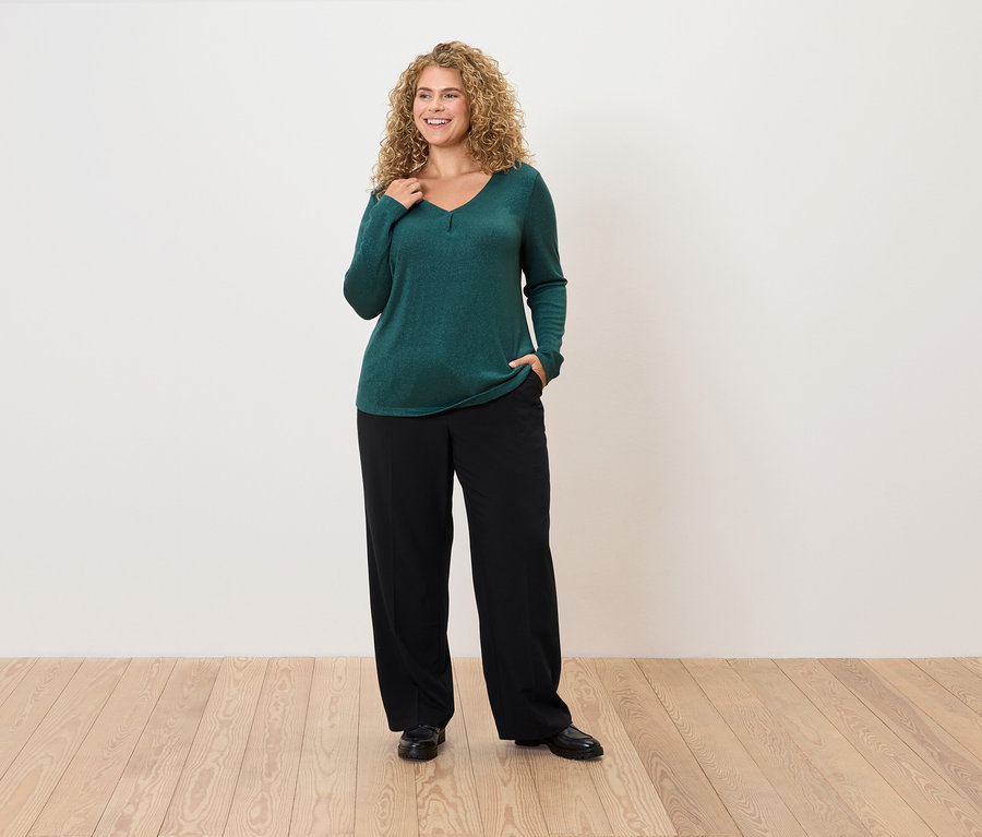 Göndör hajú nő smaragdzöld pulóverben és fekete nadrágban pózol.