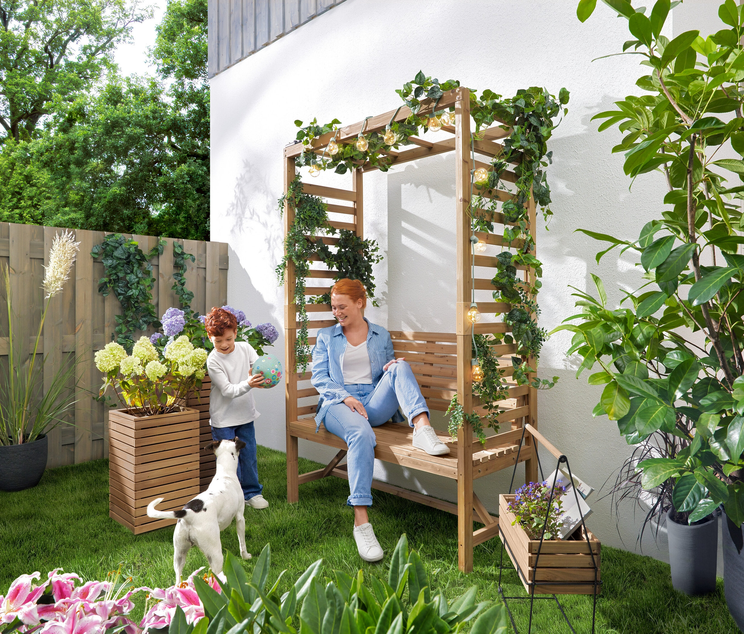 Egy nő ül egy kerti padon rózsalugassal, miközben egy gyermek labdázik. A pad fölött »Villanykörte« LED-es fényfüzér lóg. A kertben 2 db »Tinus« ültetőláda és egy »Elin« fűszernövényláda hordozókerettel található.