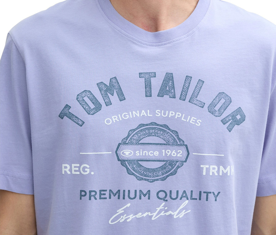 Egy világos lila "Tom Tailor" feliratú pólót viselő férfi közeli képe.