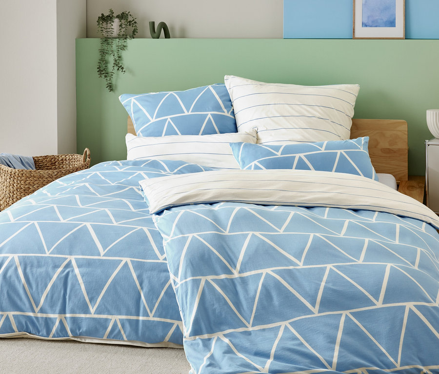 Ágy világoskék huzattal, fehér háromszögekkel és csíkos párnahuzatokkal, egy kosárkosár és egy éjjeliszekrény mellett lámpával.