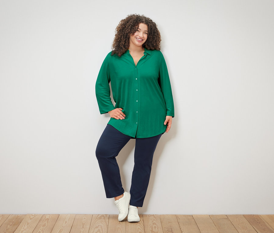 Göndör hajú nő zöld blúzban, kék nadrágban és fehér tornacipőben pózol egy fehér fal előtt.