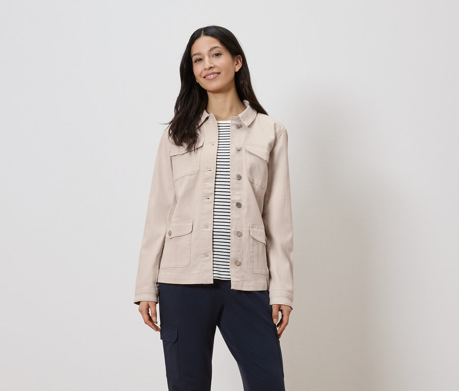 Nő visel egy bézs színű kabátot egy csíkos ing felett és sötétkék nadrágot.