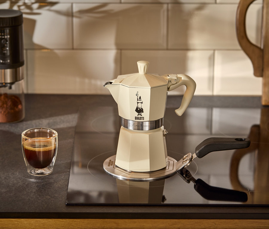 Egy krém színű Bialetti Moka Express, 190 ml-es kávéfőző áll egy Bialetti indukciós átalakítólapon a főzőlapon. A háttérben egy csésze kávé látható.