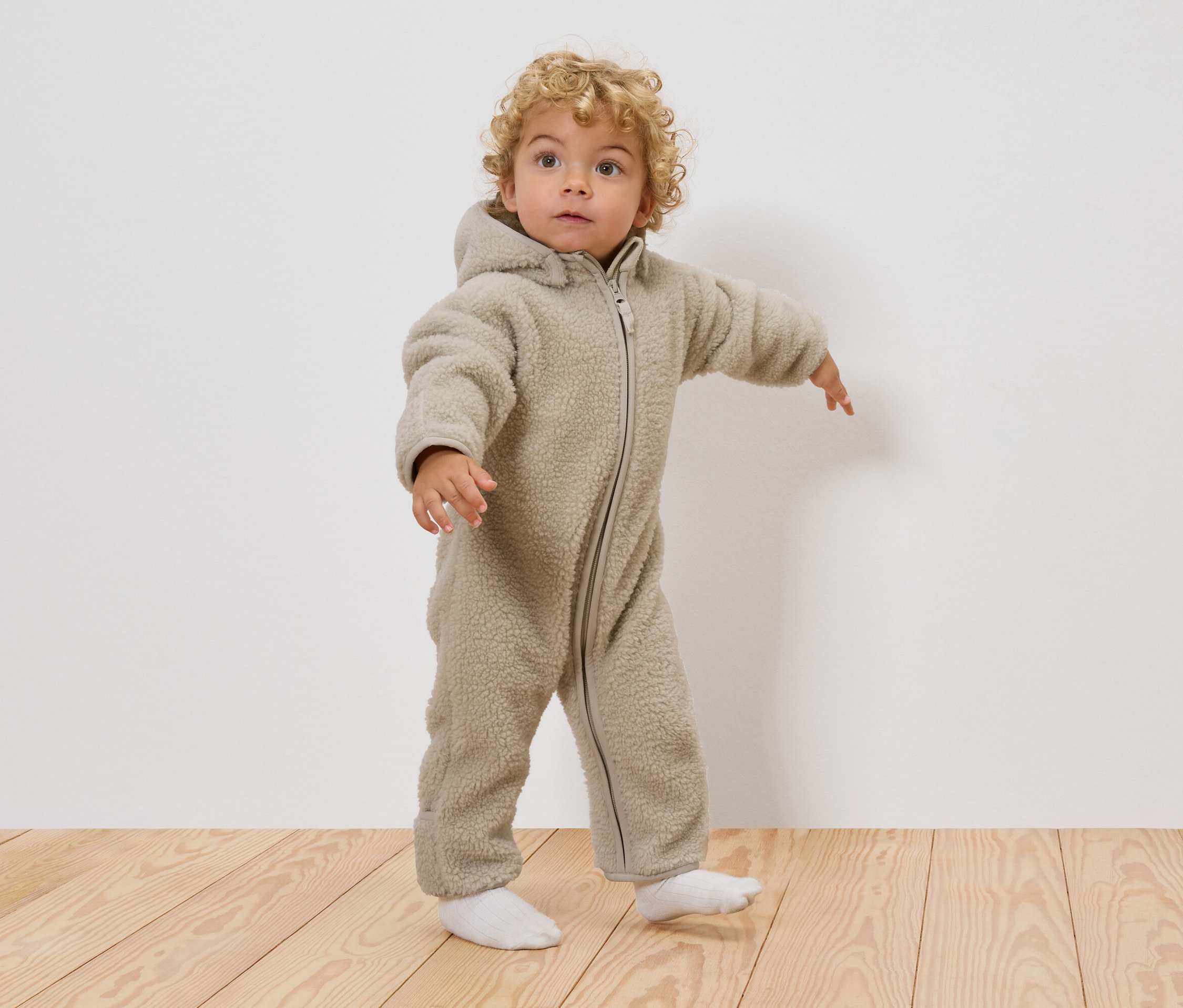 Egy göndör szőke hajú kisgyerek bézs színű, kapucnis teddy overallt és fehér zoknit visel, miközben egy fapadlón áll.