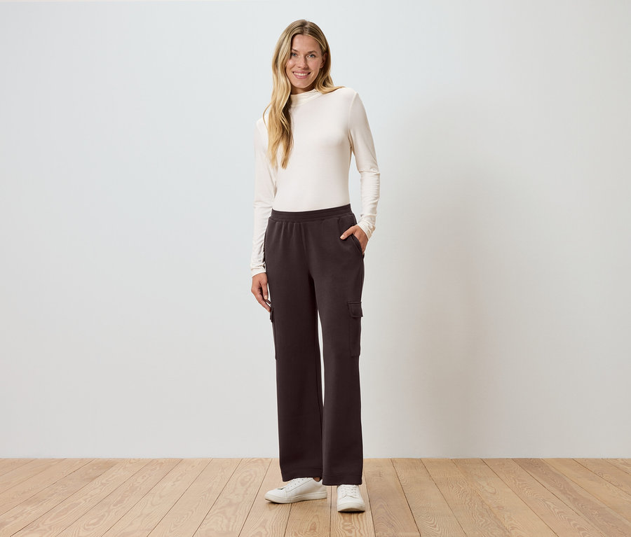 Nő áll kezével a zsebében egy fal előtt, barna cargo nadrágot visel.