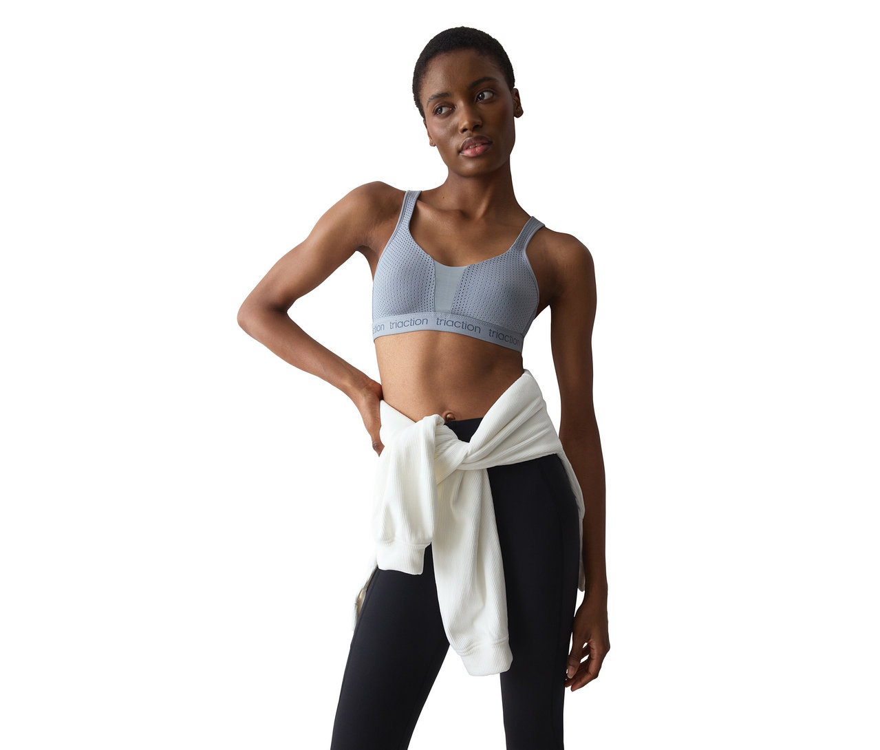 Egy rövid sötét hajú nő világosszürke sportmelltartót és fekete leggingset visel, derekán egy fehér pulóverrel. Egyik kezét csípőre téve áll, jobbra néz.