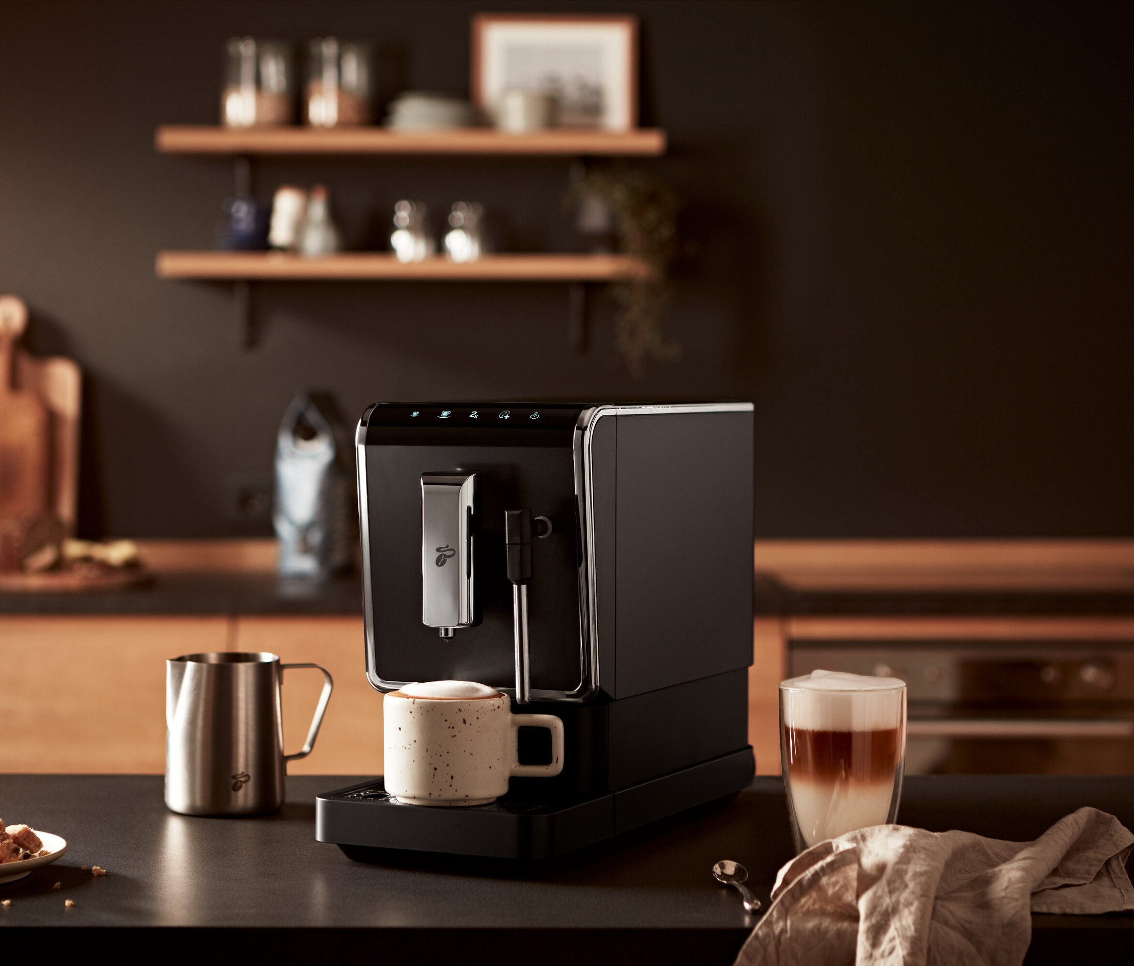 Egy asztalon egy antracit színű »Esperto Latte« Tchibo automata kávéfőző, egy csésze kávé és egy pohár latte látható.
