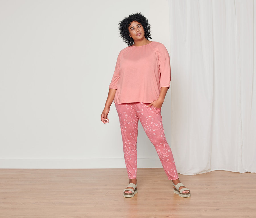 Nő pózol korall 3/4-es ujjú felsőben, korall virágos dzsörzénadrágban és bőr szandálban.