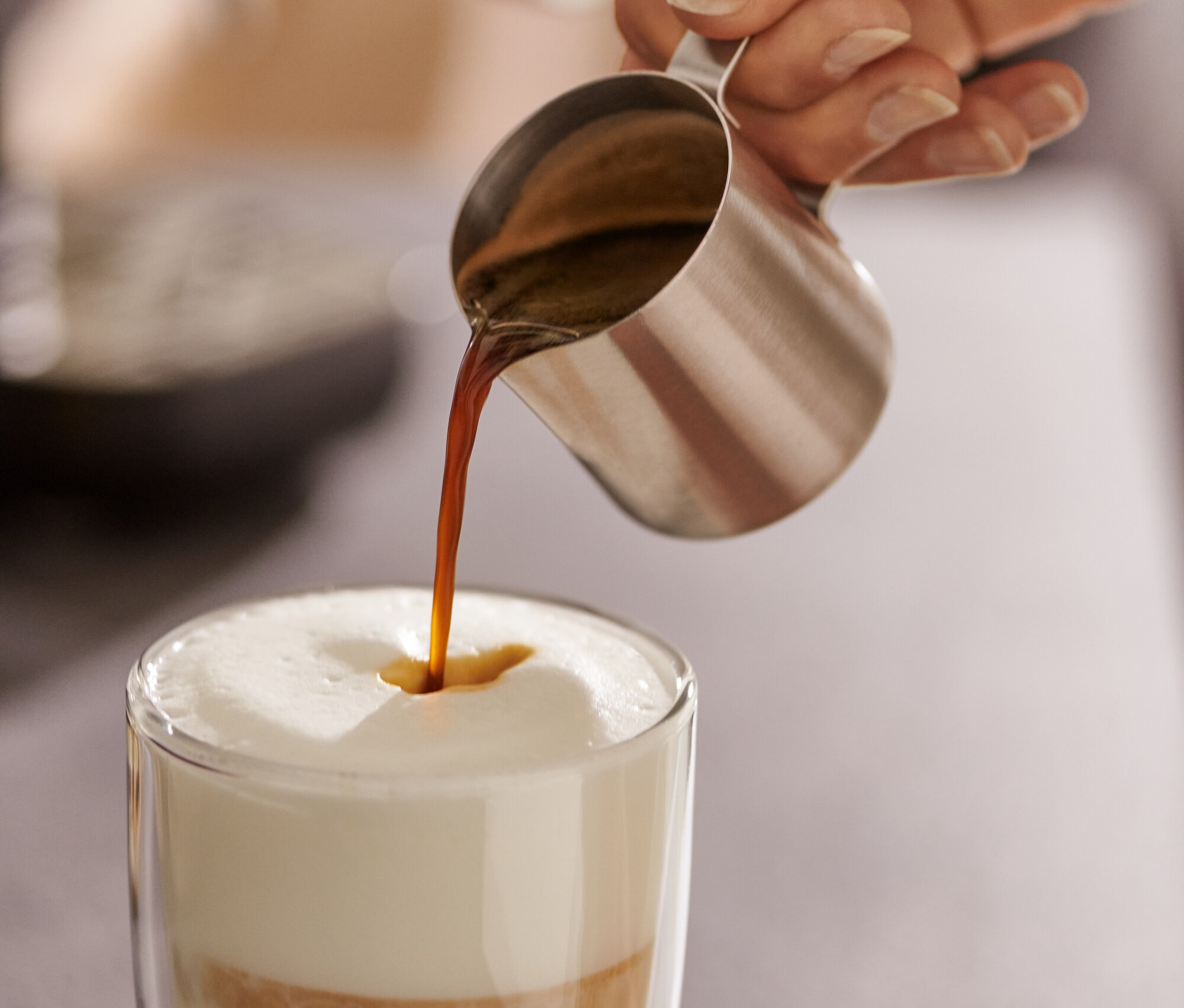 Nő eszpresszót önt egy rozsdamentes acél eszpresszó kiöntő kannából (80 ml) egy pohár tejbe.