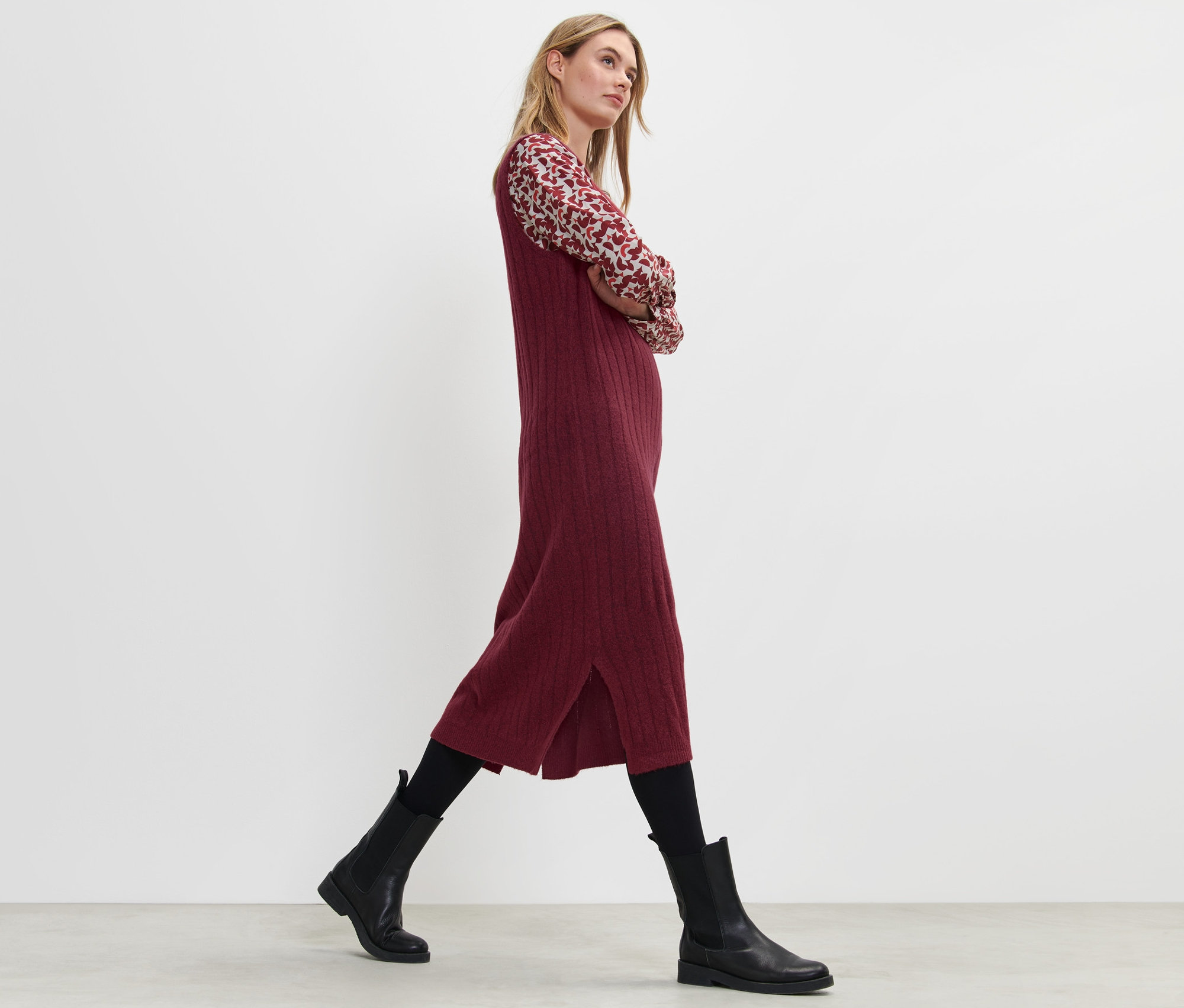 Egy szőke nő sétál, piros, bordás kötésű ruhát visel.