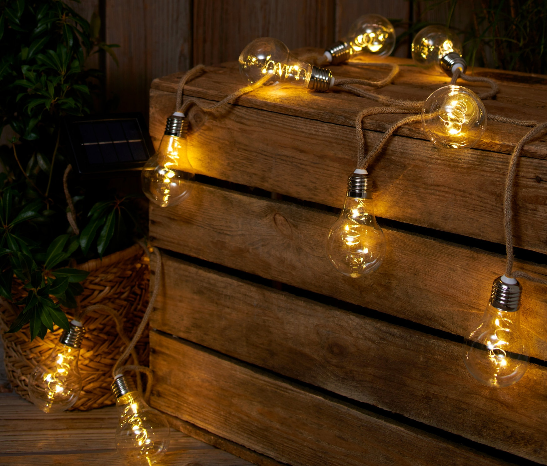 Napelemes LED-es fényfüzér juta zsinóron fekszik egy fa ládán egy növény mellett.