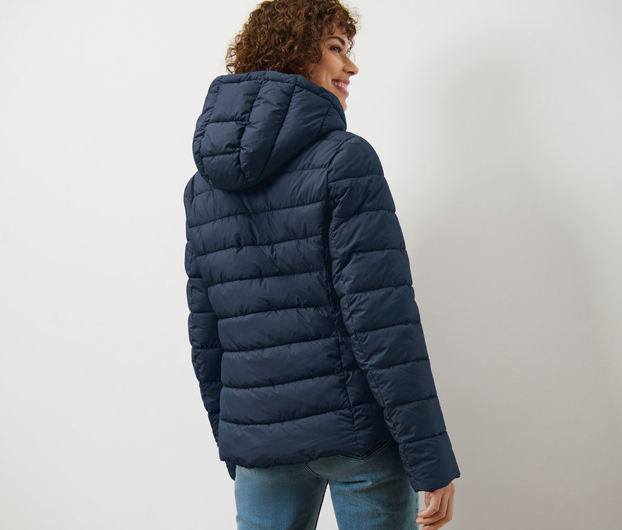 Göndör hajú nő néz a válla fölött, sötétkék, kapucnis steppelt dzsekit visel.