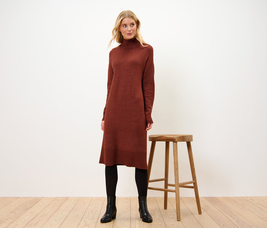 Nő áll egy fa szék mellett, és gyapjú tartalmú kötött ruhát visel.