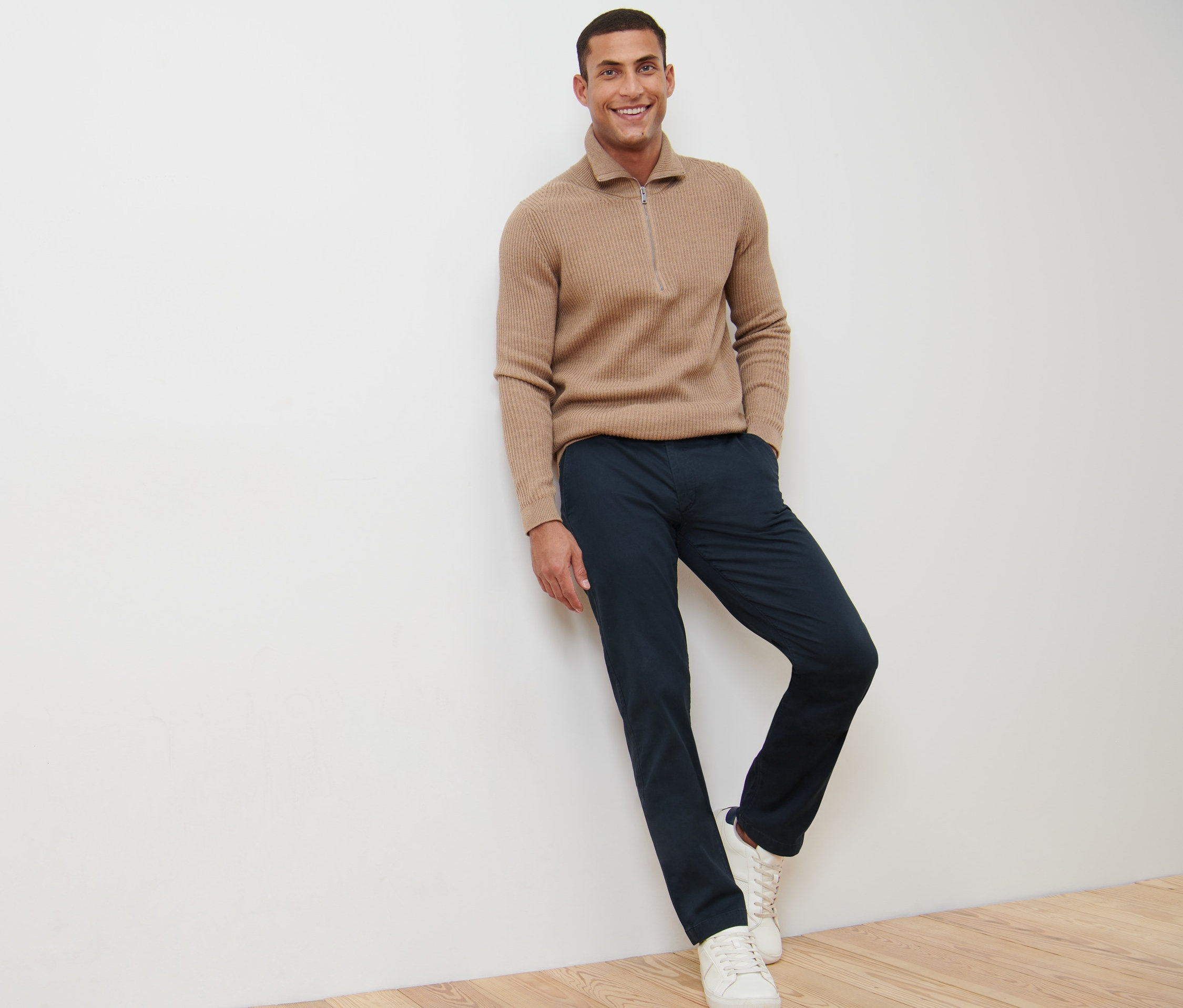 Egy mosolygó férfi a falnak támaszkodik, tengerészkék chino nadrágot visel.