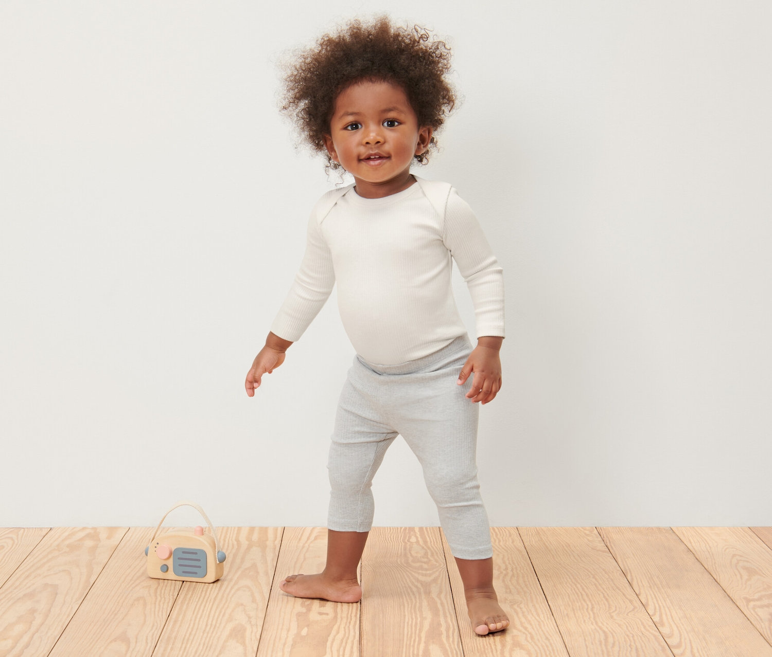 Egy kisgyerek áll a fapadlón, és 3 kisgyerek hosszú ujjú body, törtfehér színű bodyt és szürke leggingset visel.