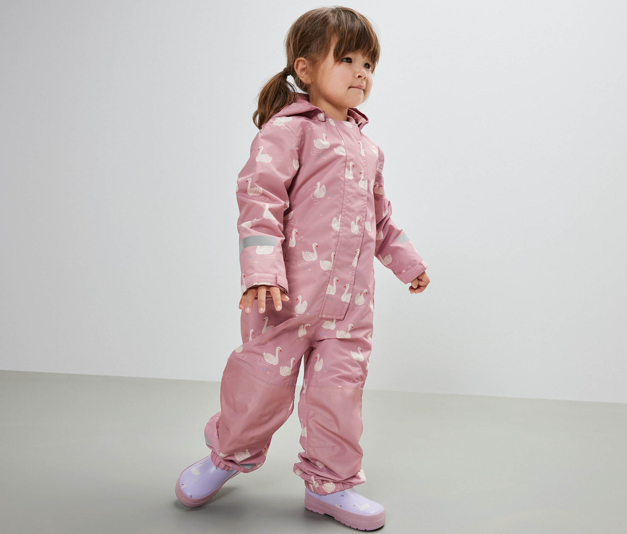 Kislány eső elleni kezeslábasban, hattyús. Lila gumicsizmát visel.