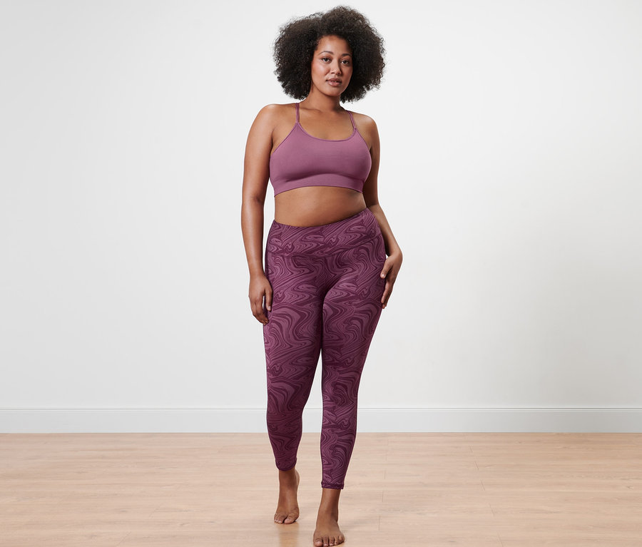 Nő pózol mályva színű női seamless bustier sportmelltartóban és mintás női 7/8-os sportnadrágban.