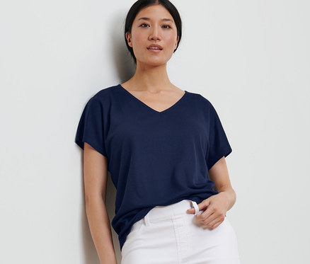 Egy falnak dőlő nő sötétkék strukturált felsőt és fehér nadrágot visel.