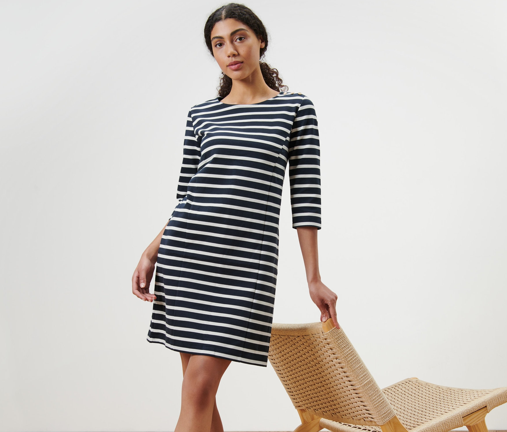 Nő pózol egy csíkos női lezser ruhában egy vesszőfotel mellett.