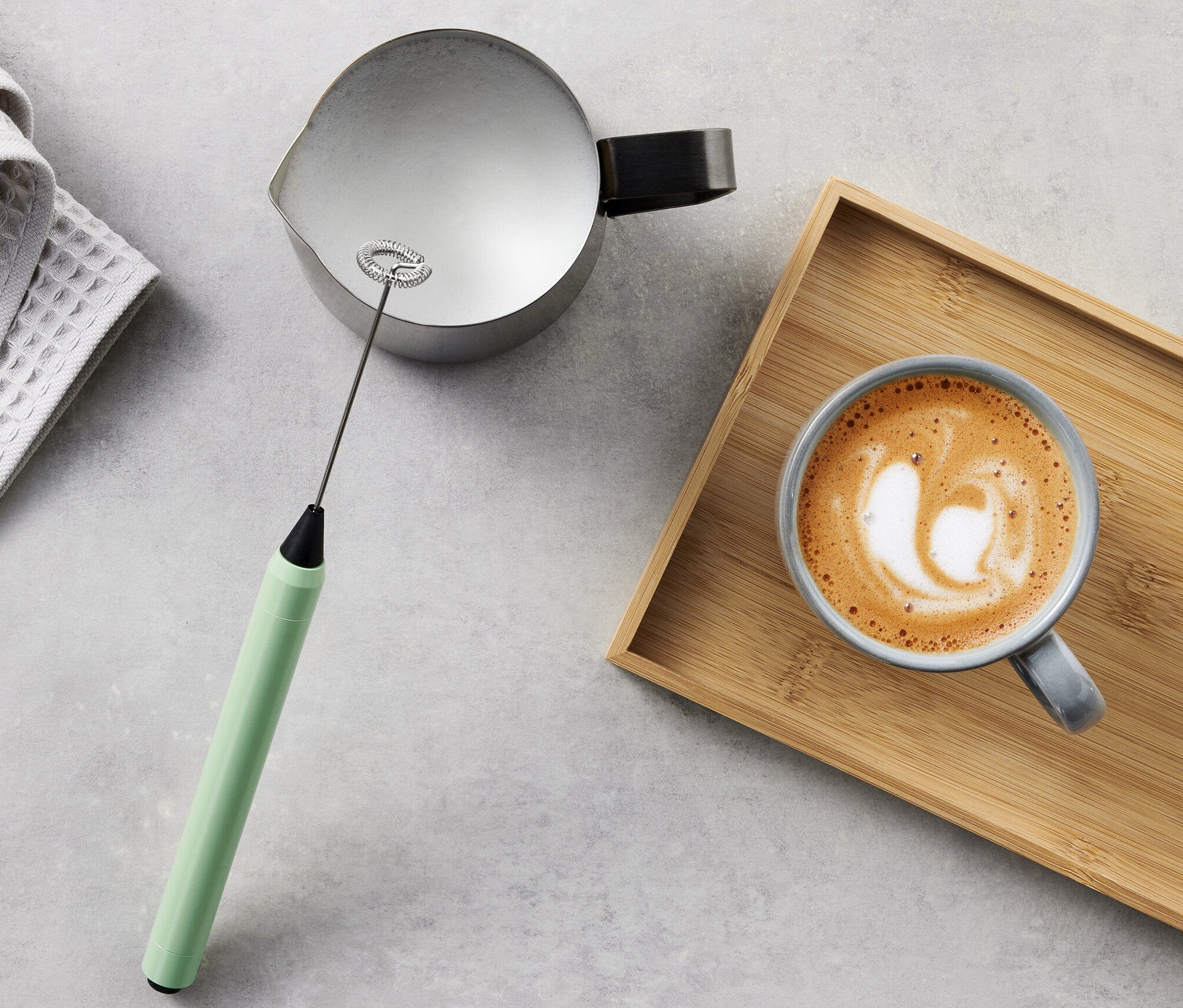 Szürke törlőkendő, kiöntő, kézi tejhabosító és tejeskávé fa tálcán.