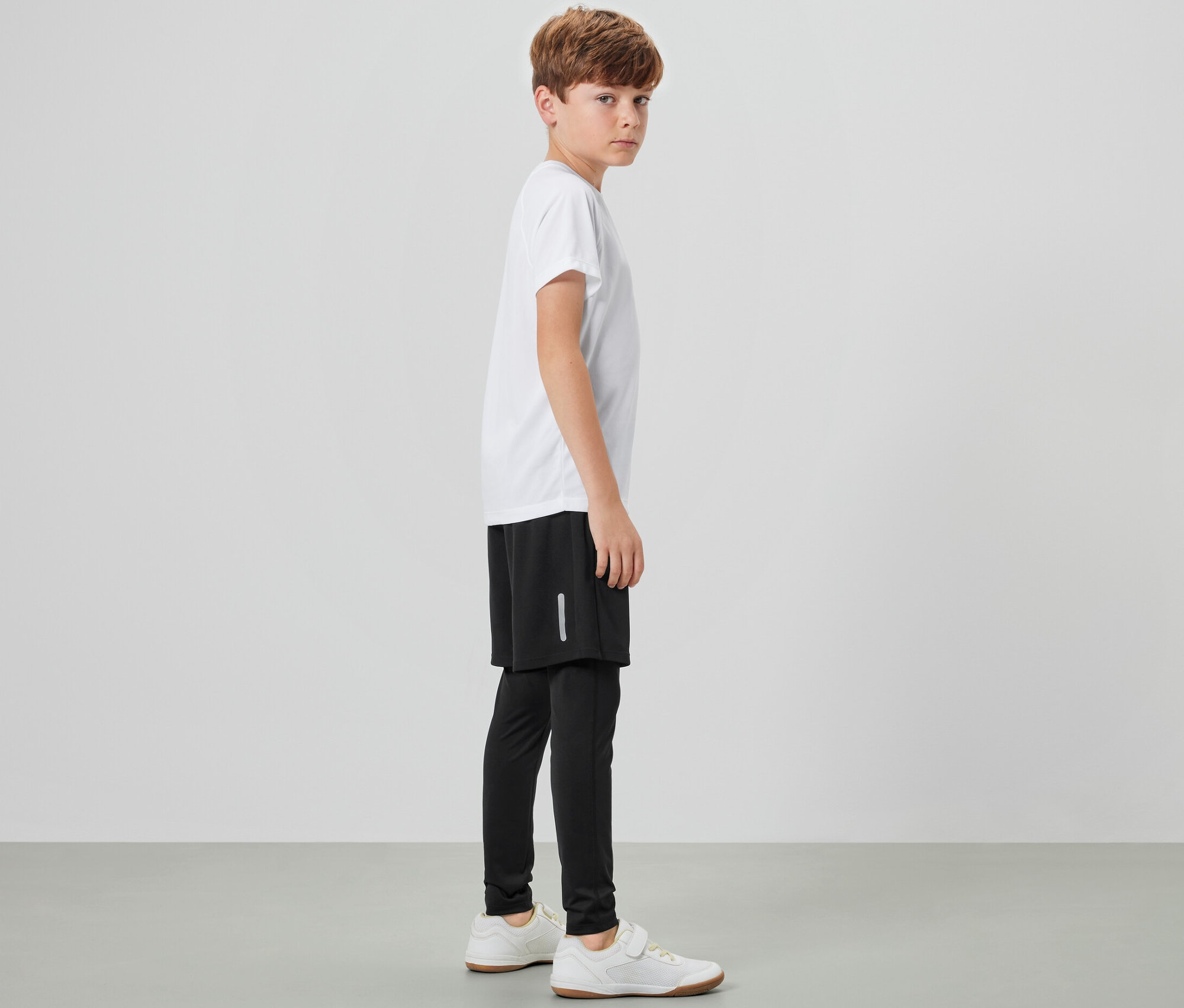 Egy oldalt álló fiú gyerek kétrétegű sportnadrágot, fehér pólót és tornacipőt visel.